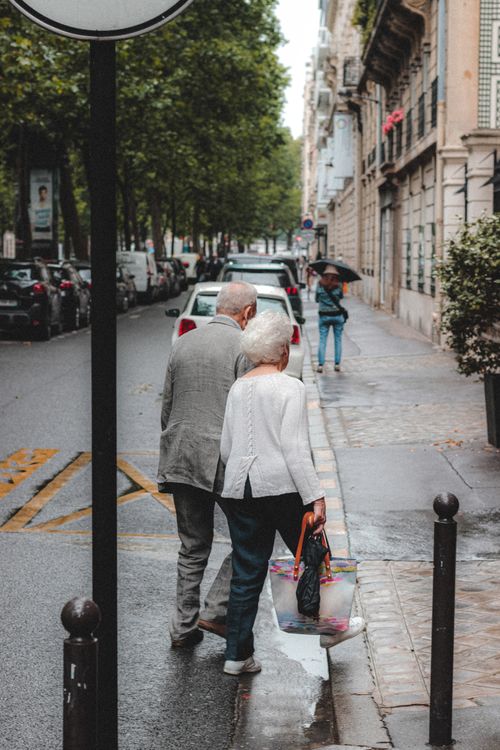 Free Full body back view senior couple in formal wear walking across asphalt road surrounded by buildings and parked automobiles Stock Photo