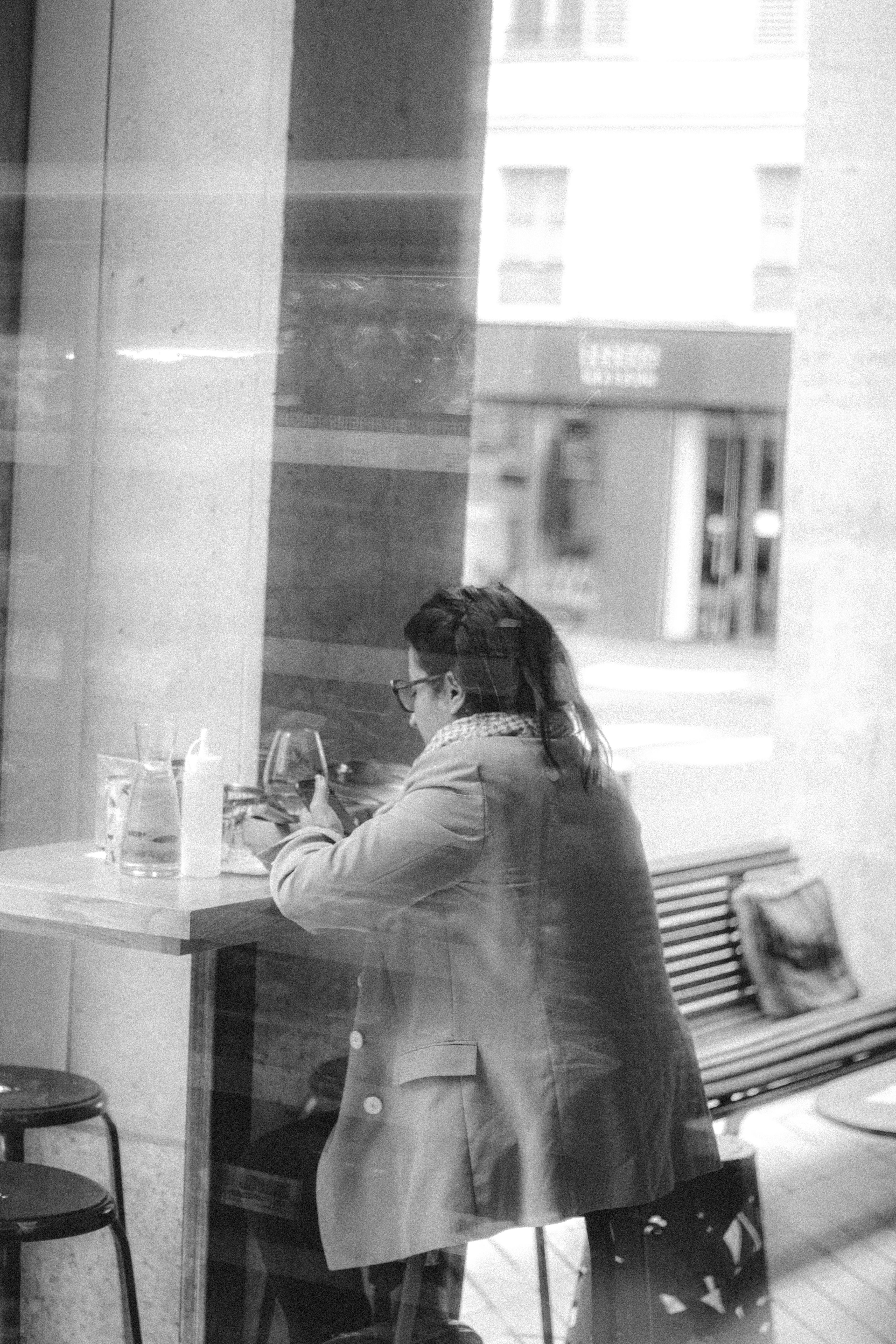 Black and white through glass back view of unrecognizable female text messaging on cellphone at table in cafeteria
