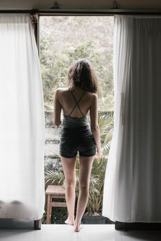 Back view of young slim unrecognizable female traveler enjoying view of tropical forest while chilling on terrace of bungalow