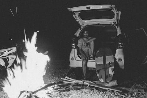 Black and white bearded young man sitting in car trunk near surfboard and looking at bonfire at night