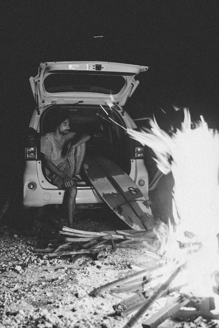 Pensive Young Man Sitting In Car Trunk Near Surfboard Against Campfire