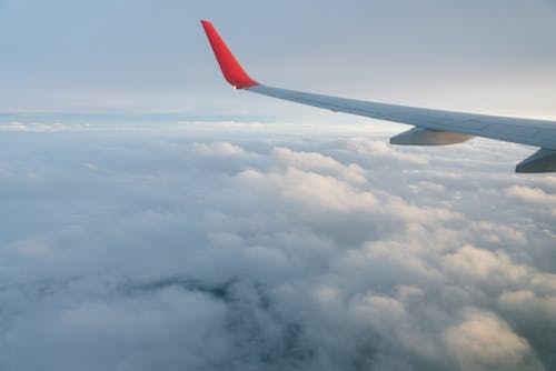 Through window of red and white wing of aircraft flying over fluffy clouds in endless blue sky