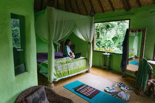 Free Full body female resting on comfy bed with netbook while spending day in cozy green room of rural cottage located amidst exotic rainforest Stock Photo