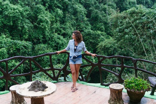 Full body calm female in denim leaning on house balcony railing with arms outstretched and enjoying tropical jungle view in exotic resort