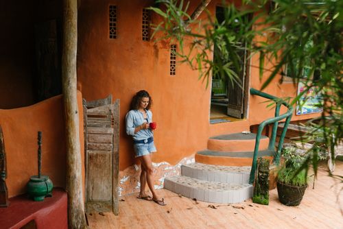 Free Full body brunette wearing denim clothes with cup of hot drink leaning on rural cottage wall while visiting exotic tropical country Stock Photo