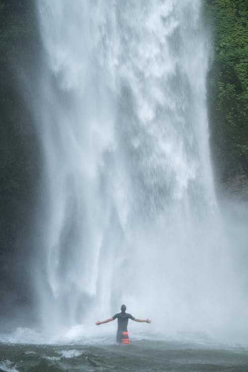 Back view faceless male tourist in black shirt standing with outstretched arms in lake near waterfall and admiring spectacular view