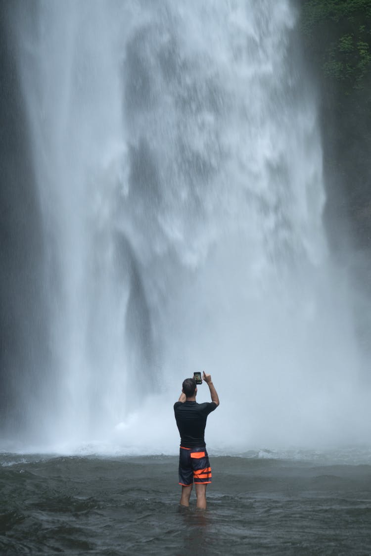 Male Traveler Taking Photo Of Majestic Waterfall On Smartphone