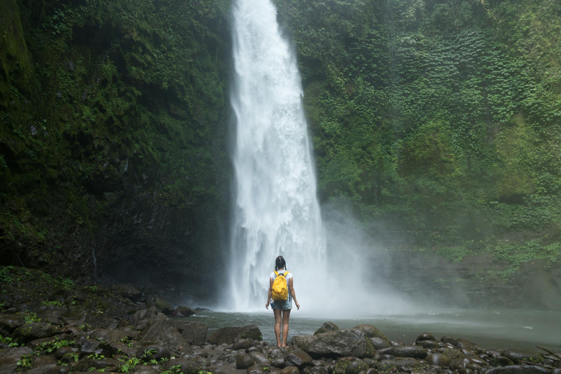 Back view unrecognizable female backpacker standing on stones near spectacular waterfall while traveling in exotic country