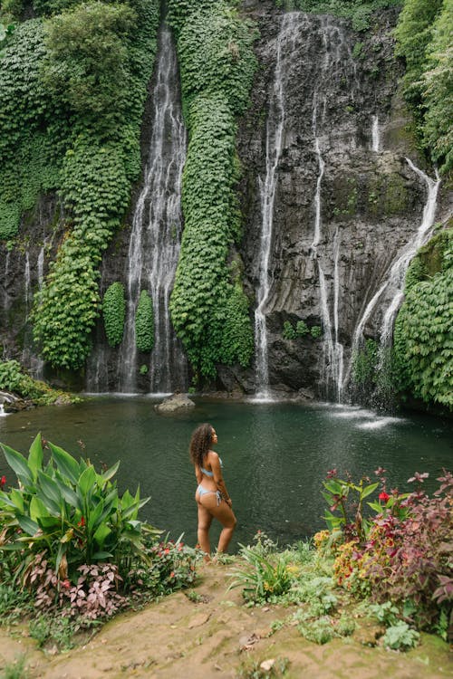 From above of unrecognizable young female tourist in swimwear standing in front of pond and waterfall in rainforest
