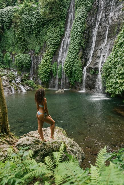 Side view of faceless gorgeous curly haired female tourist in bikini standing on rough stone on lake shore against powerful waterfall