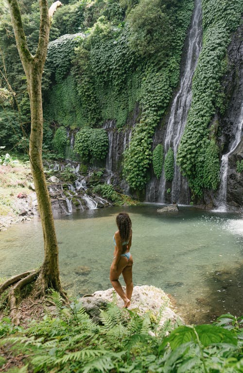 Back view of unrecognizable young fit barefooted female in bikini resting in canyon near crystal clean lake and waterfall