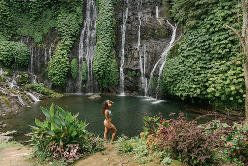 High angle of peaceful young woman in bikini touching hair while standing between tropical plants against waterfall flowing into pond