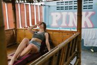 Young happy female in crop top and shorts laughing cheerfully covering face with hand while relaxing on bamboo daybed with thatched canopy