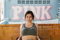 Young confident good looking female in melange gray sports top with tied up hair sitting on bamboo bench on balcony and looking at camera