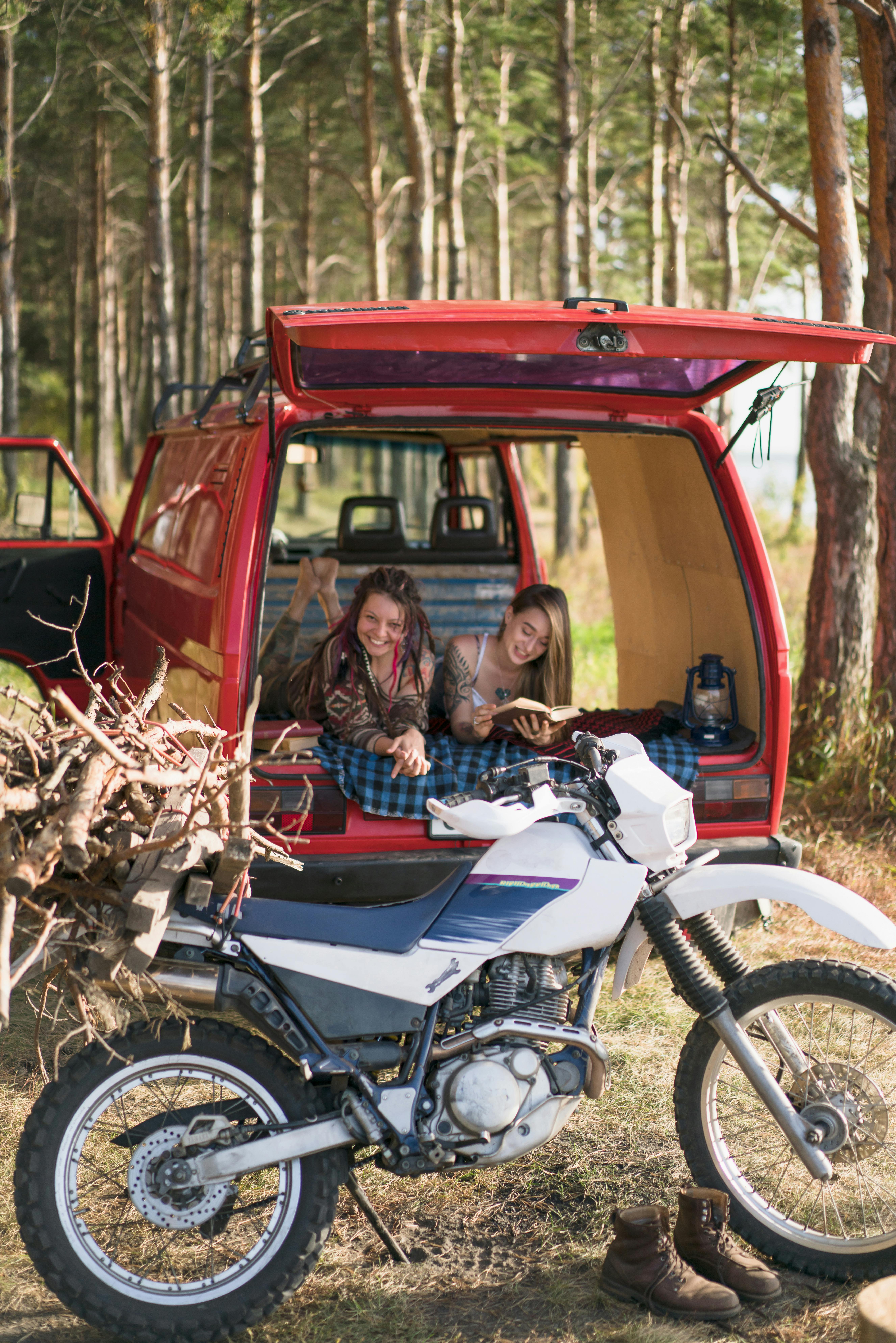 Two women enjoying a forest adventure, relaxing with a van and motorcycle setup for a perfect outdoor experience.