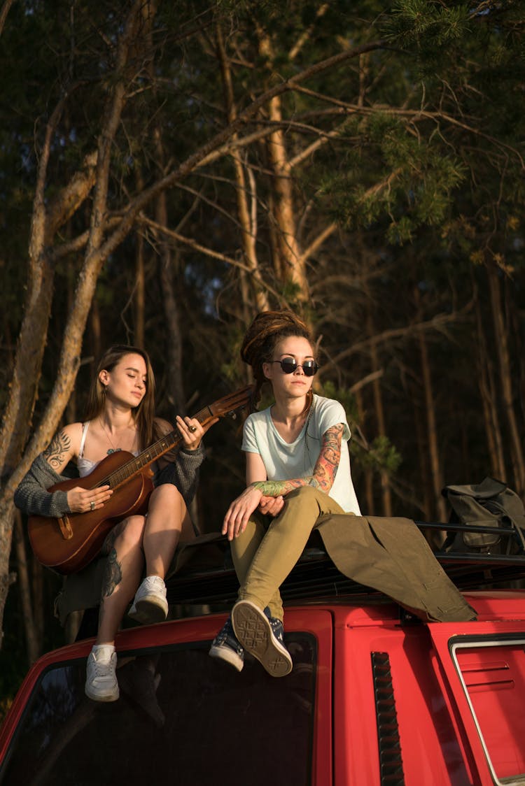 Woman Sitting On Car Roof On Camping And Playing Guitar