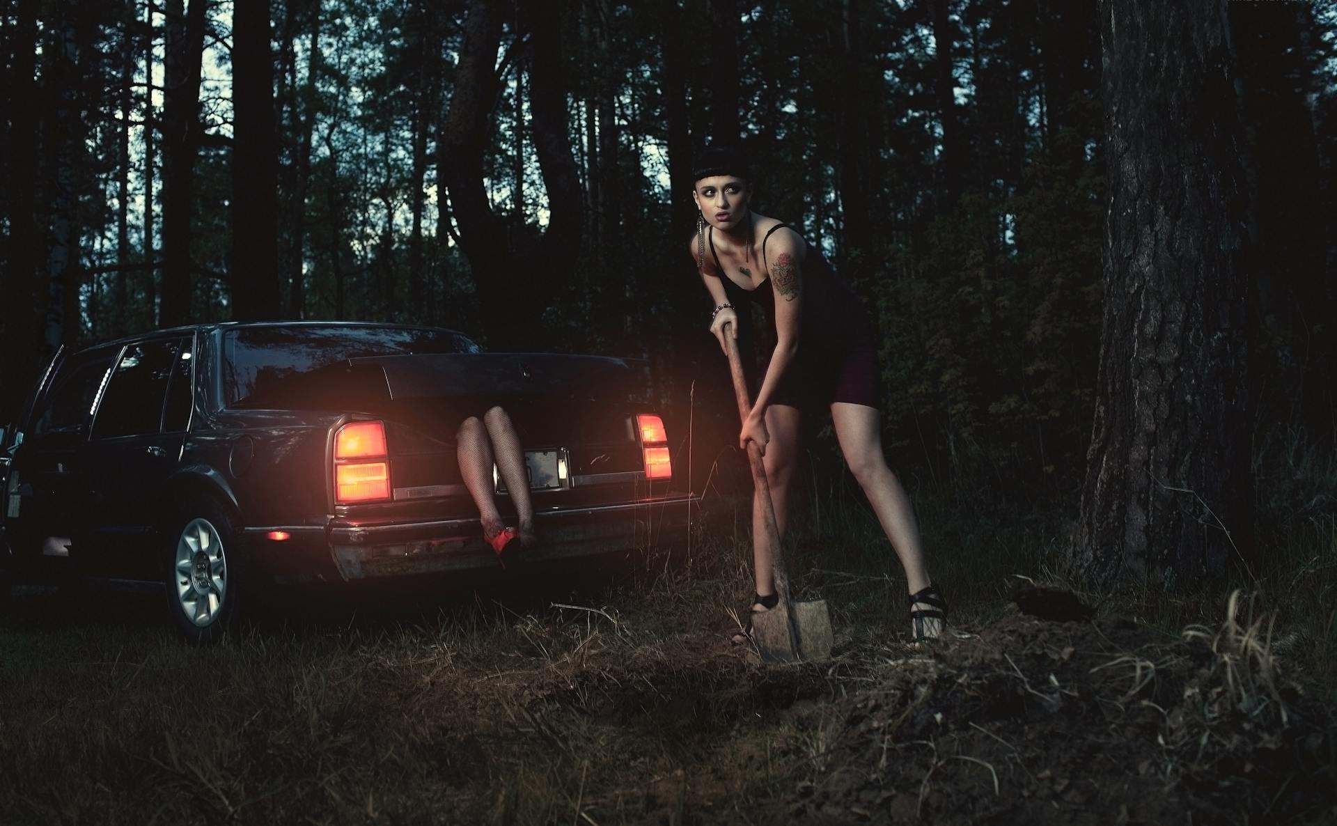 A woman digs in the forest next to a car with eerie overtones.