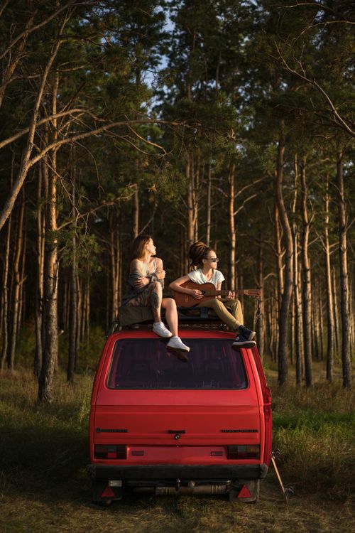 Free Couple sitting on van roof in forest, playing guitar and soaking in the nature at sunset. Stock Photo