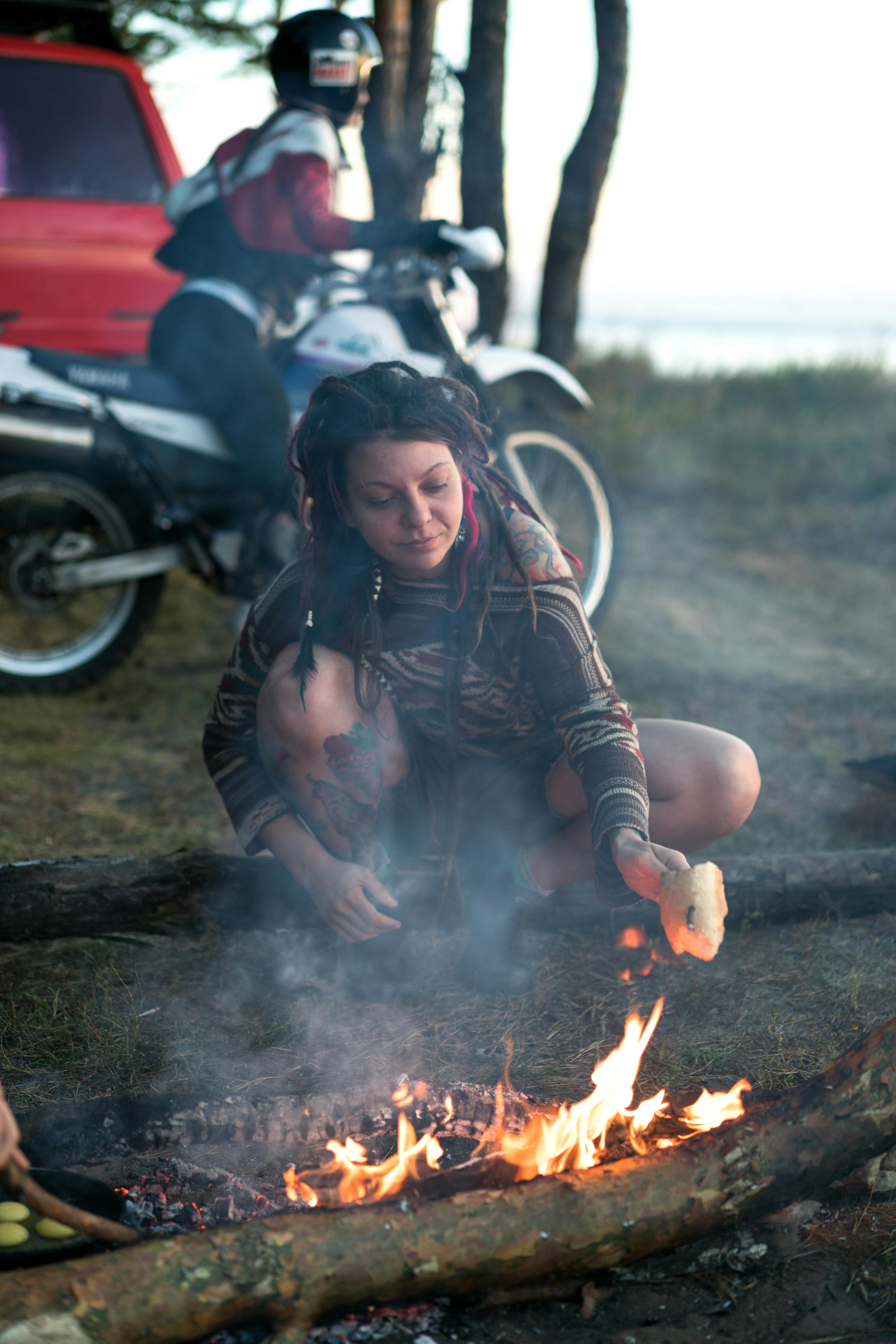 Woman crouches by campfire cooking food, with motorcycle nearby, outdoors setting.