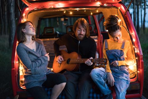 Free Three friends play guitar and relax in a van with cozy lights in the woods. Stock Photo