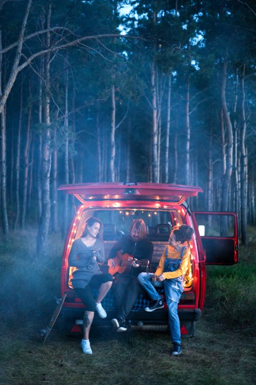 Free Three young adults playing guitar and enjoying a campfire by a red van in a forest setting at twilight. Stock Photo