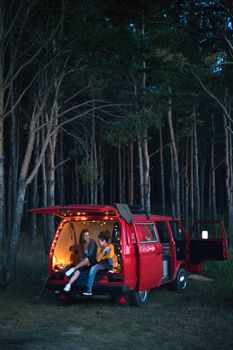 Two women enjoy a serene summer evening in a van parked in a tranquil forest setting.
