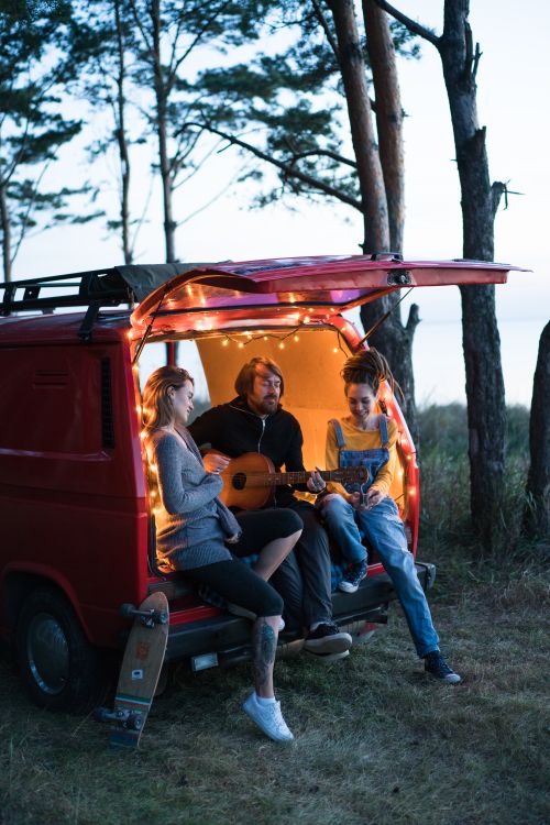 Free A group of friends relaxing and playing guitar by a camper van in the woods at dusk. Stock Photo