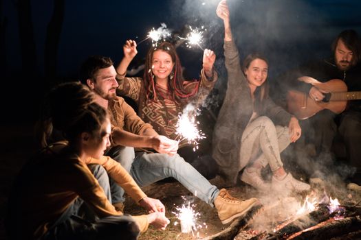 Group of friends sitting by a bonfire, holding sparklers and enjoying a night under the stars.