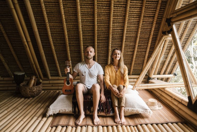 A couple sitting indoors in a bamboo villa, holding a ukulele, enjoying a rustic lifestyle.