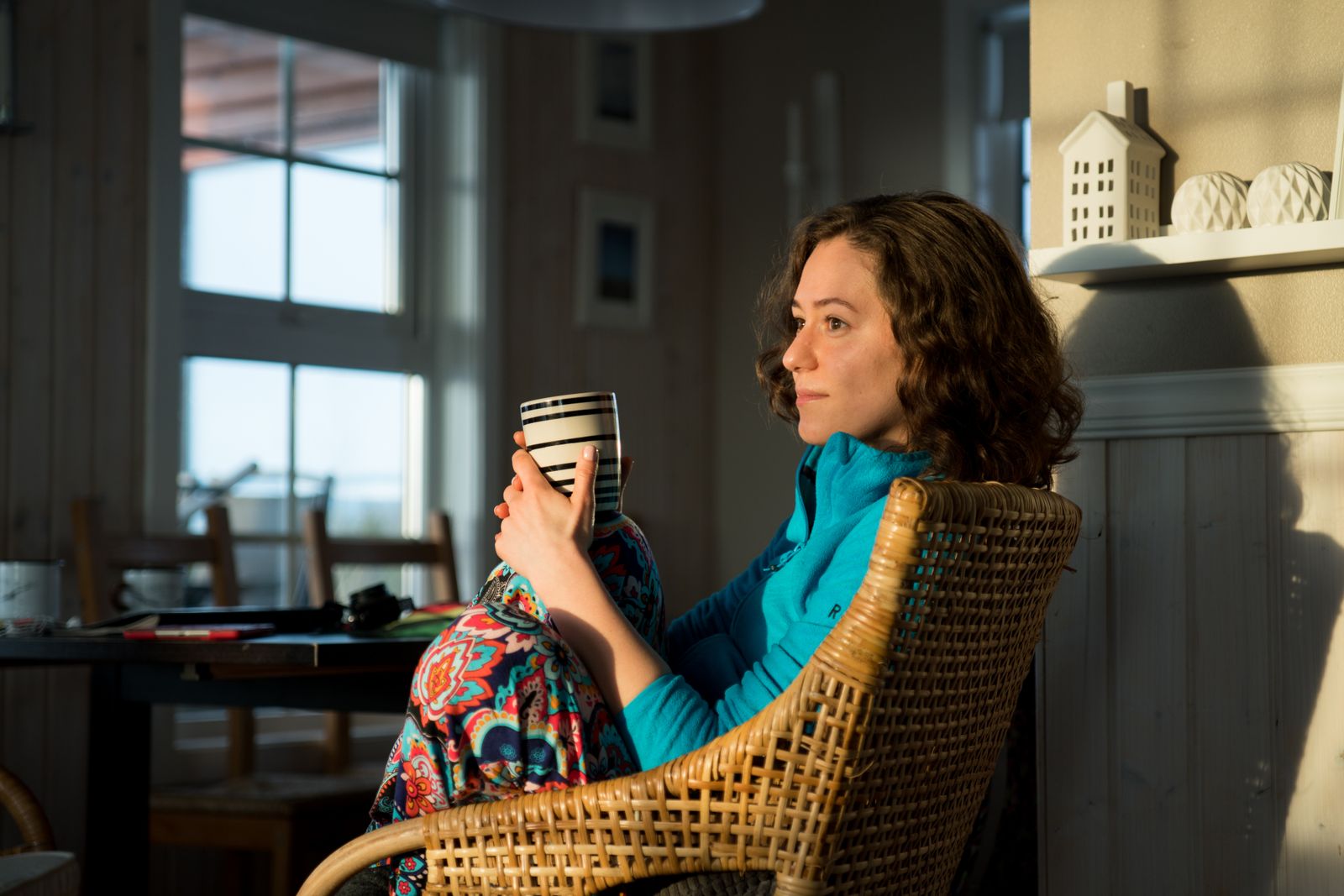 Woman with curly hair sits in a wicker chair holding a striped mug in warm sunlight