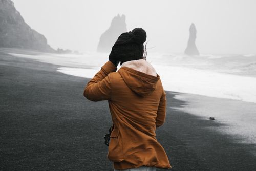 Free A woman walks on a windy beach with dramatic sea stacks in the distance, showcasing an adventurous coastal landscape. Stock Photo