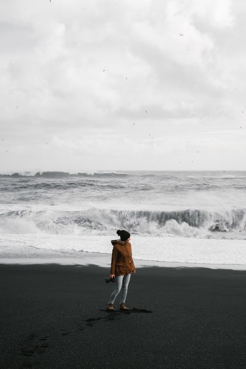Free A woman stands on a remote black sand beach with waves crashing under an overcast sky. Stock Photo