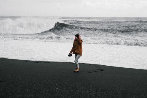 Free A woman in a brown jacket walks on a black sand beach, holding a camera with waves crashing in the background. Stock Photo