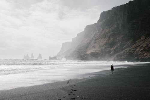Free A lone figure walks along a misty, dramatic Icelandic beach with towering cliffs and crashing waves. Stock Photo