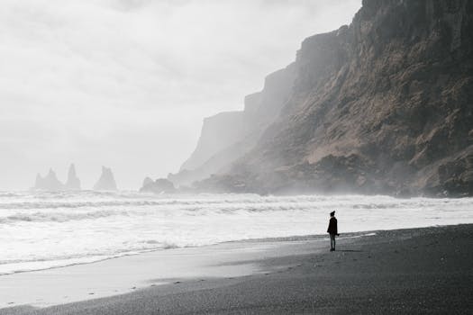 A lone person stands on a foggy beach in Iceland, facing the misty ocean and towering rock formations.