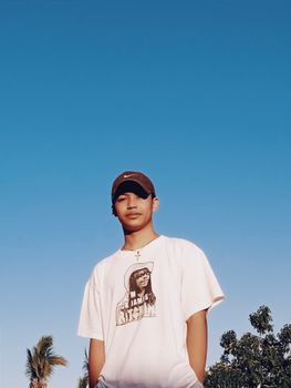 From below of confident ethnic adolescent in cool t shirt standing against cloudless blue sky