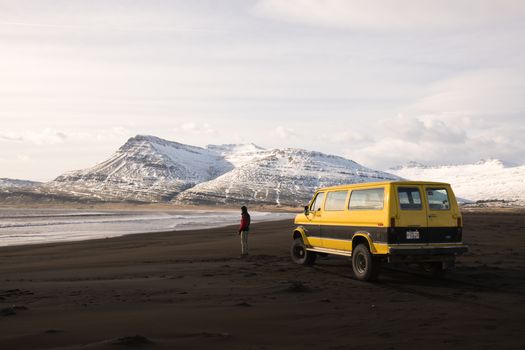 A man stands by a yellow van on a sandy shore with snowy mountains in the background.
