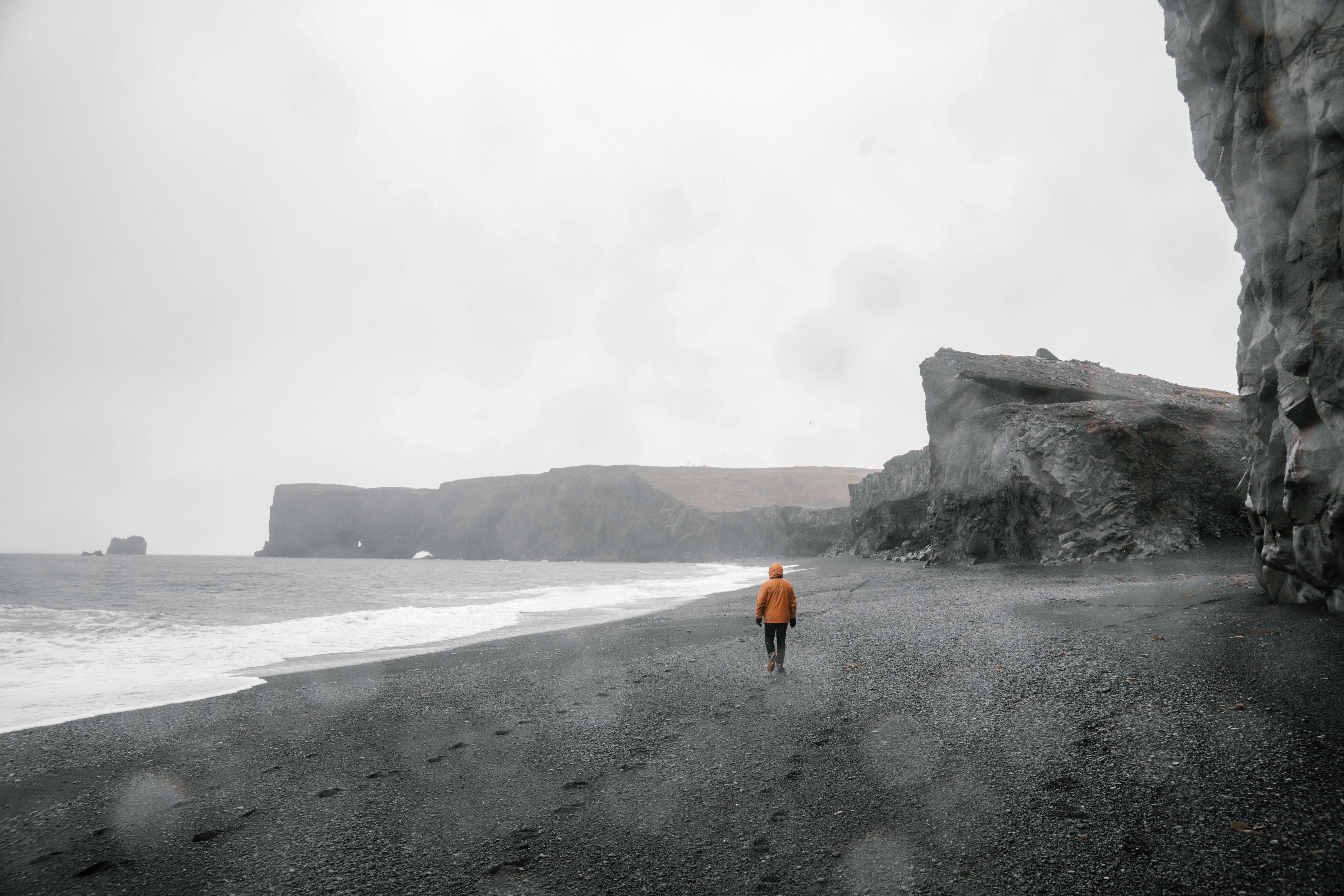 A person in a raincoat walks on a rugged black sand beach with towering cliffs and overcast skies.
