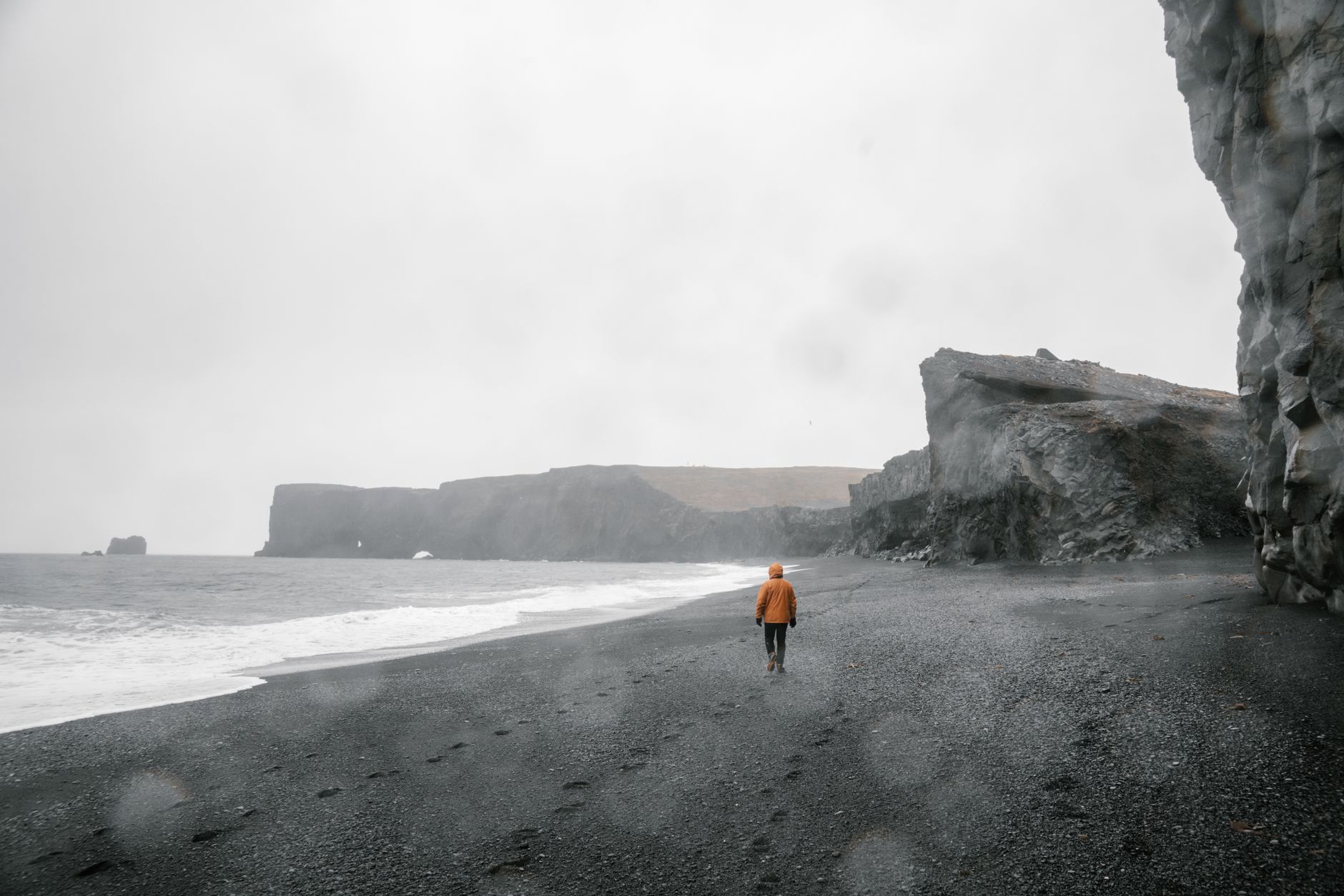 A person in a raincoat walks on a rugged black sand beach with towering cliffs and overcast skies.