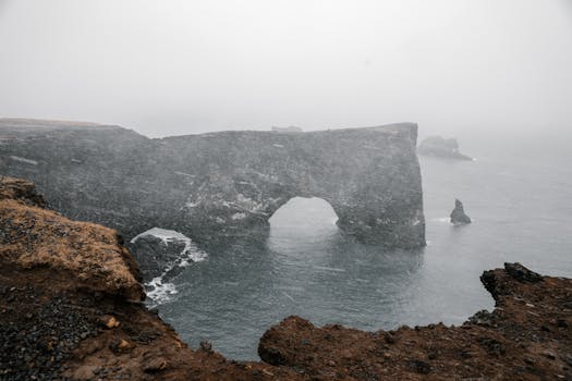 Dramatic view of a sea cliff arch over stormy waters during a snowstorm, creating a moody atmosphere.