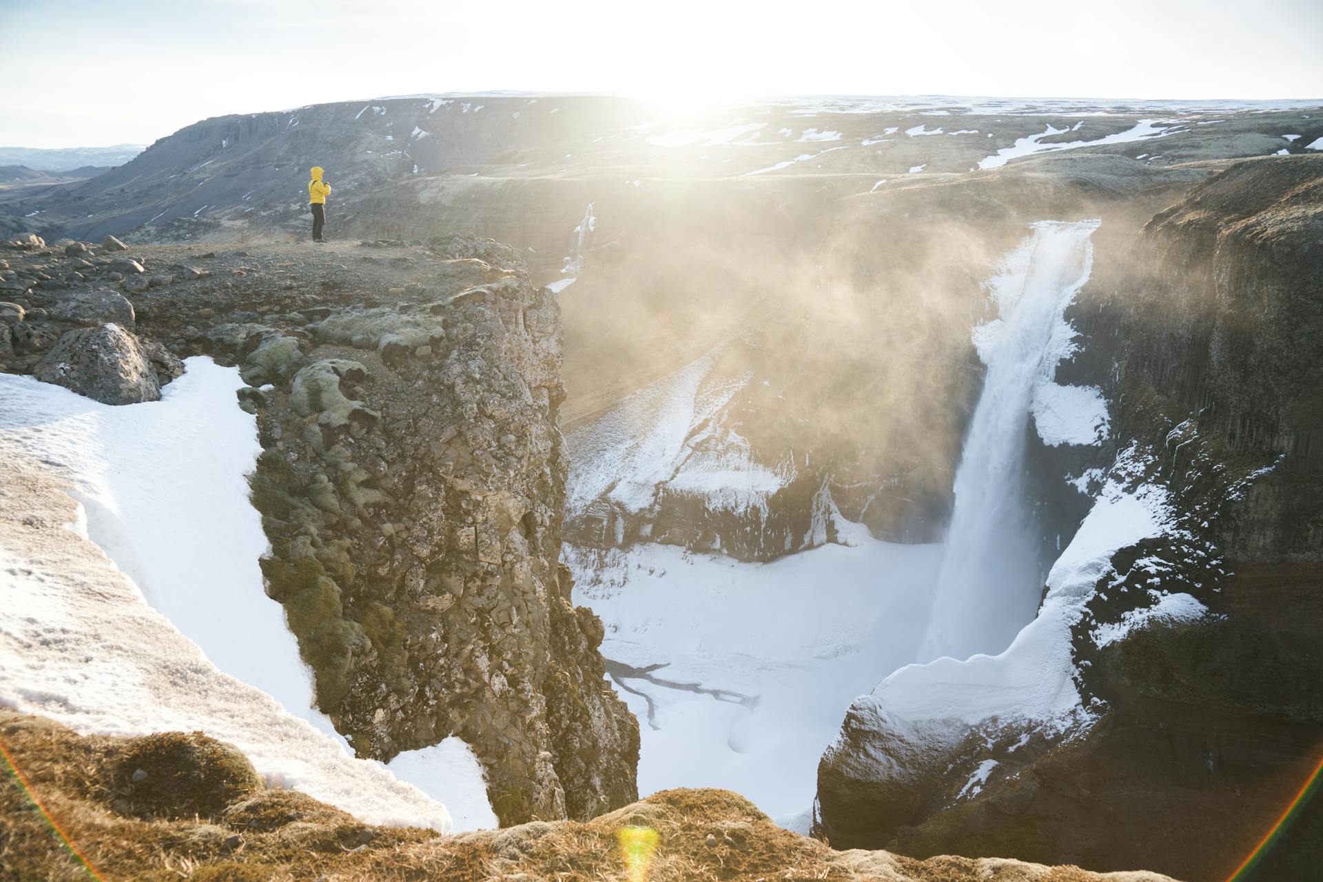 A lone person in a yellow jacket stands on a cliff edge with a waterfall cascading into the snowy landscape below.