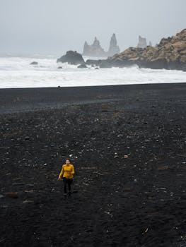 A woman in a yellow jacket walks on a misty black sand beach with rock formations.