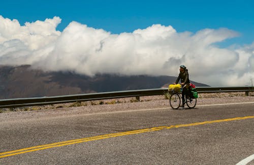 Unrecognizable guy in warm clothes and helmet riding bicycle on asphalt road with flowers in basket and admiring amazing scenery of clouds over mountains