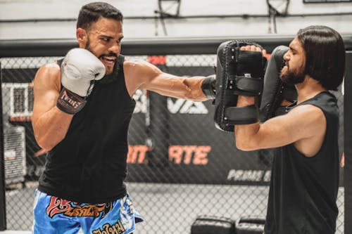 Serious determined ethnic male fighter in sportswear and gloves performing punches on boxing paws during workout with trainer in ring