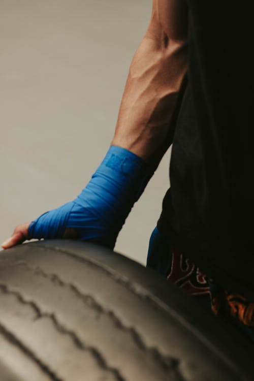 Free Crop unrecognizable muscular sportsman with blue hand wrap carrying heavy tire during functional training in gym Stock Photo