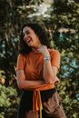 Cheerful young female with dark curly hair in casual outfit laughing while standing in green park near trees