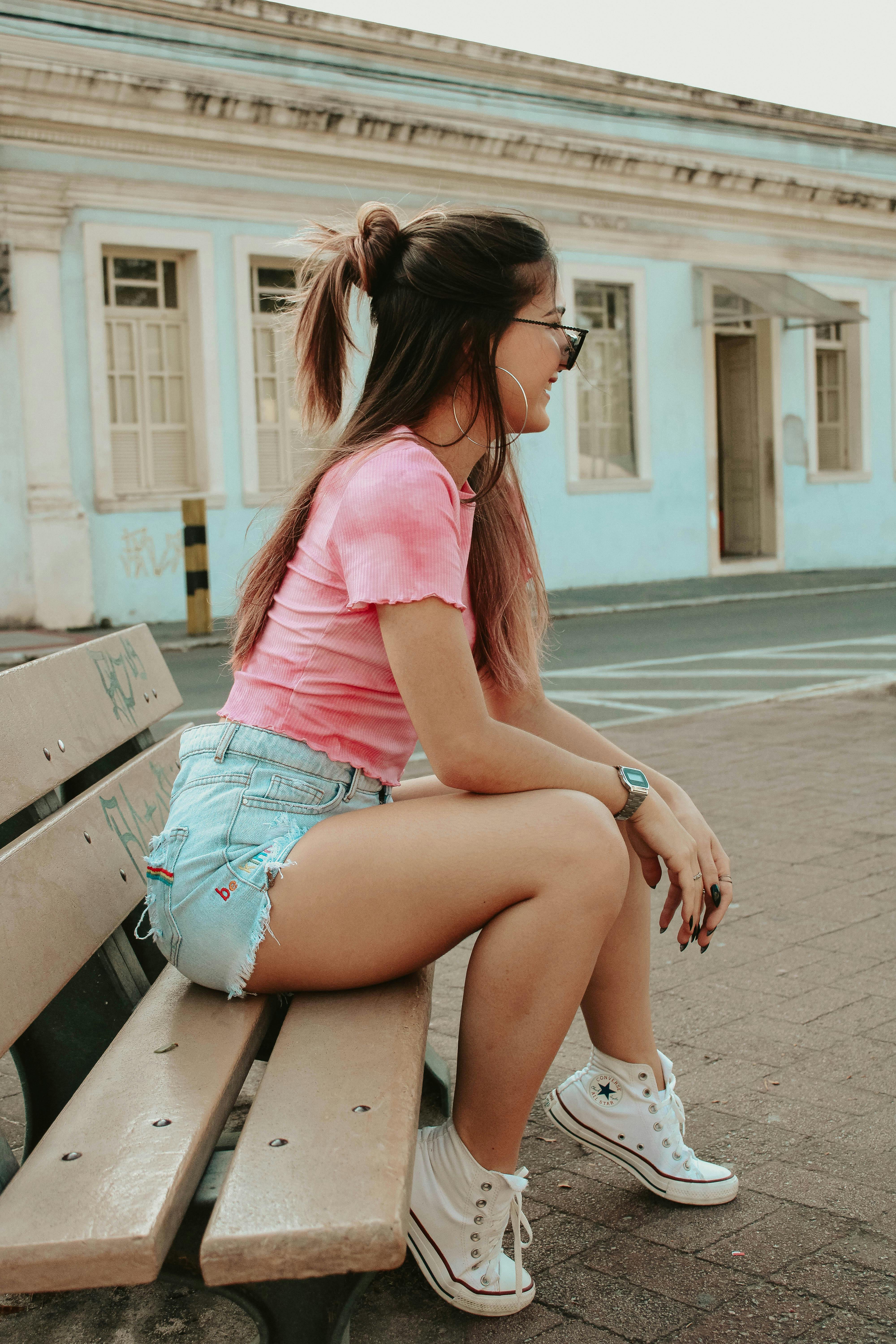 Side view of happy young stylish female millennial in trendy sneakers and sunglasses relaxing on wooden bench on city street and looking away