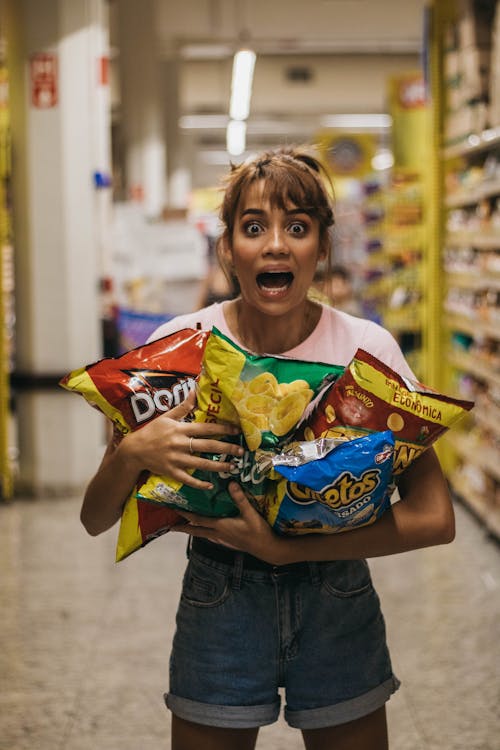 Funny young female in casual clothes screaming while carrying various packs of potato chips in supermarket