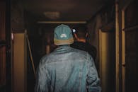 一名男子身穿牛仔夹克、头戴帽子，在光线昏暗的走廊里留下背影