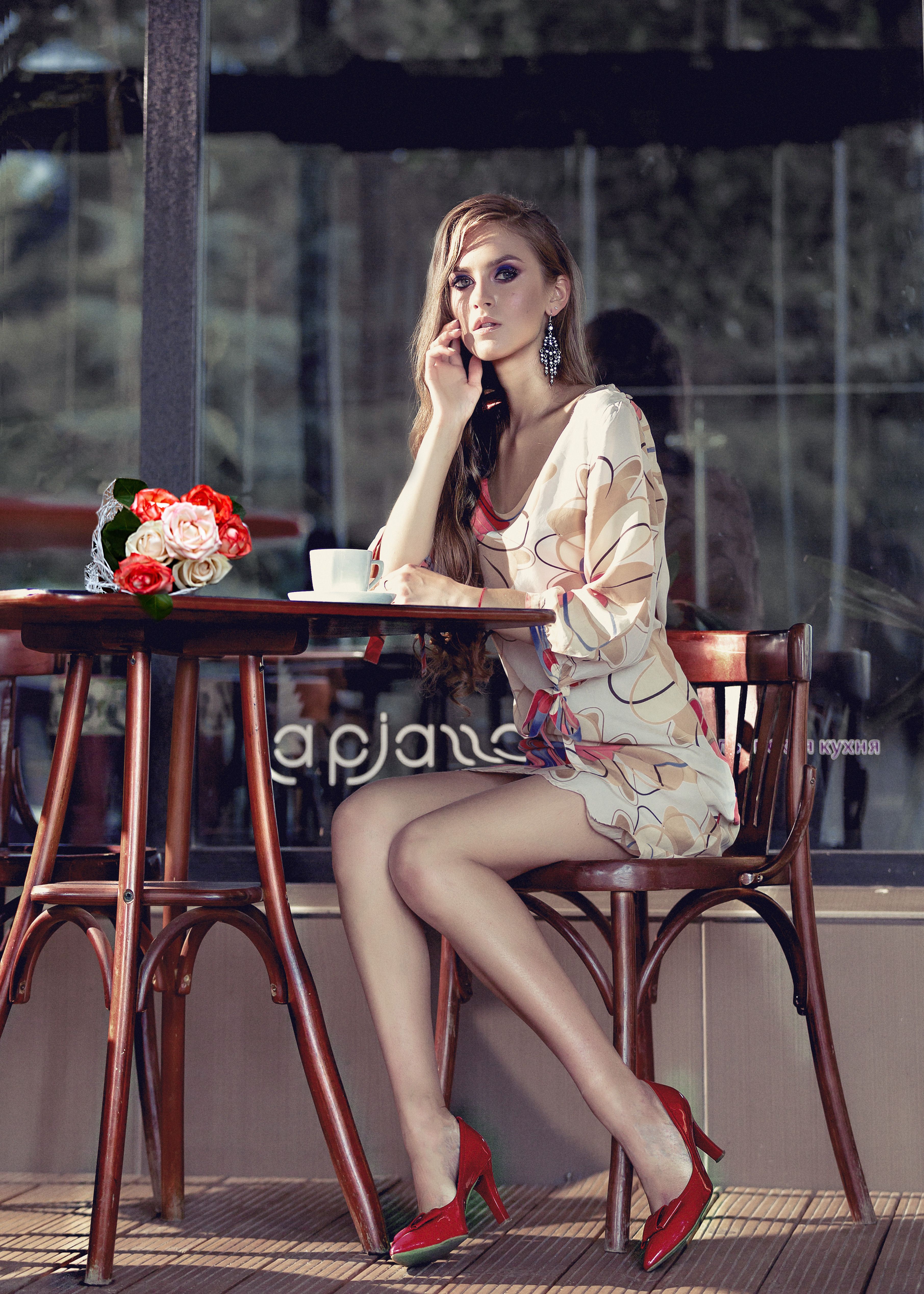 Side view of self assured elegant female in high heels and dress sitting at table in street cafe with coffee cup and bunch of flowers